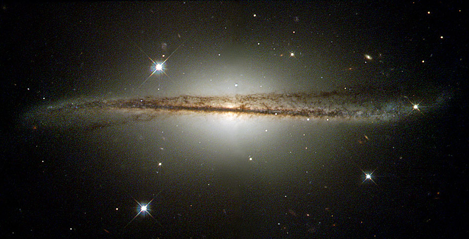 Die diffus wirkende Galaxie im Bild ist von einer sehr markanten dunklen Staubbahn überzogen. Die Staubbahn verläuft leicht gekrümmt.