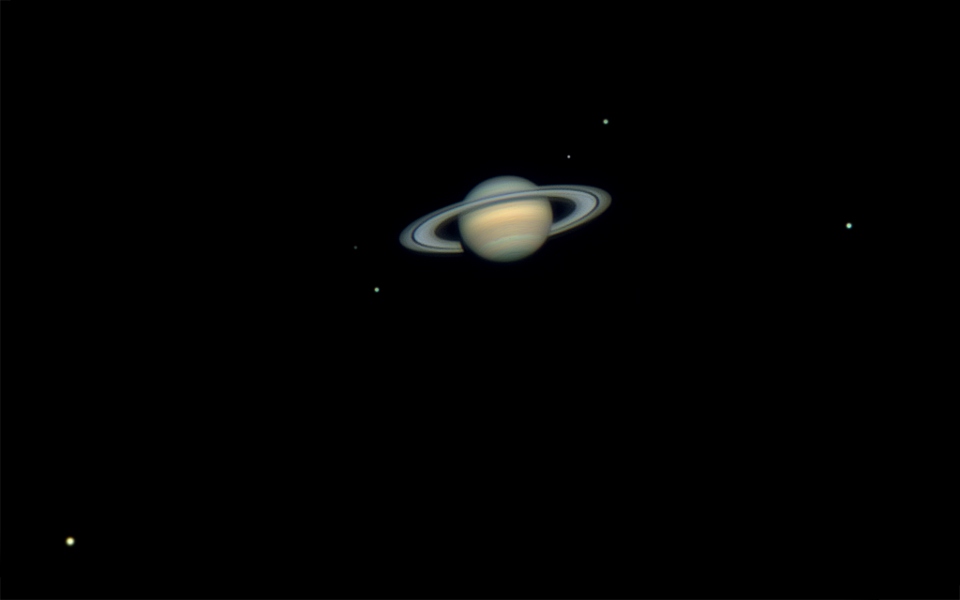 Der Ringplanet Saturn ist von unten zu sehen. Seine Oberfläche ist von ockerfarbenen Wolkenbändern überzogen. Im Bild sind seine sechs hellsten Monde verteilt.