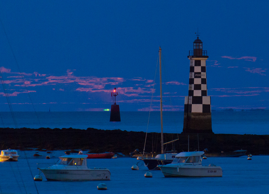 Die Landschaft ist in dunkelblaues Licht geraucht, vorne schwimmen Boote. Rechts steht ein Turm mit auffälligem Schachbrettmuster, der Tourelle de la Perdrix. In der Mitte auf dem Meer schwimmt eine Boje mit rotem Leuchtfeuer, dahinter ist ein grüner Blitz über dem Horizont zu sehen, es ist die Spitze des aufgehenden MOndes.