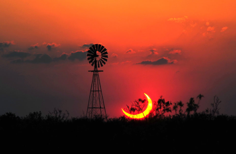 Hinter einem Wald ragt die Silhouette einer Windmühle auf, rechts daneben leuchtet die Sichel der Sonne bei der partiellen Sonnenfinsternis am Sonntagabend am rot gefärbten Himmel.