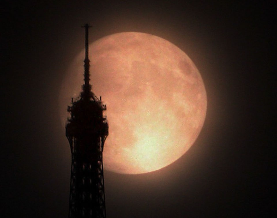 Der gelb-bräunlich leuchtende Mond mit dunklen Flecken leuchtet hinter der Spitze des Eiffelturms, die als Silhouette zu sehen ist.