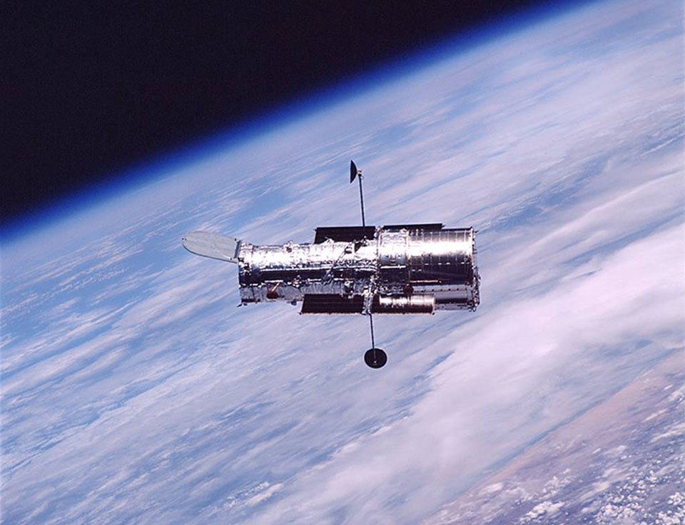 Über der Erde mit Wolken und Ozeanen schwebt das Weltraumteleskop Hubble. Links oben geht die Atmosphäre mit einem blauen Rand in die Schwärze des Weltraums über.