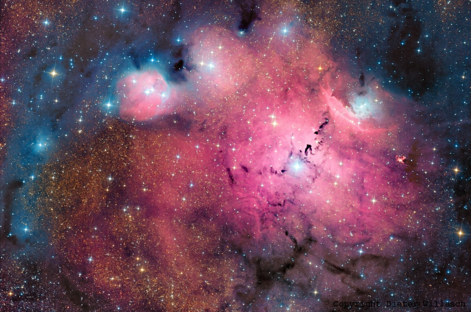 Ein magentafarbener Nebel füllt fast das ganze Bild, links oben gesäumt von blauen Reflexionsnebeln und durchzogen von einigen dunklen Staubranken. Im Bild leuchten einige markante helle Sterne, dazwischen sind Sterne wie Glitzer verteilt.