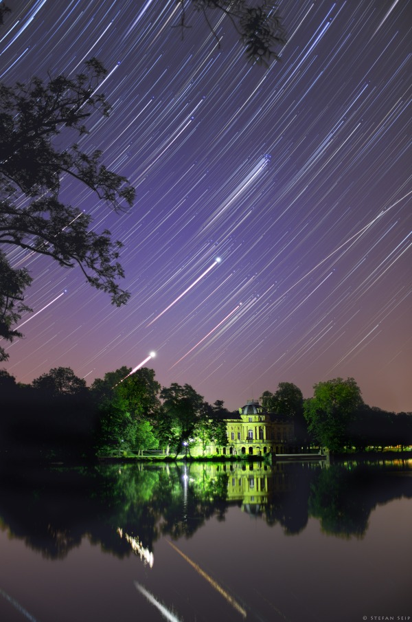 An einem spiegelglatten See steht ein grün beleuchtetes Schloss. Am Himmel ziehen Strichspuren, die sich teilweise im ruhigen See spiegeln.