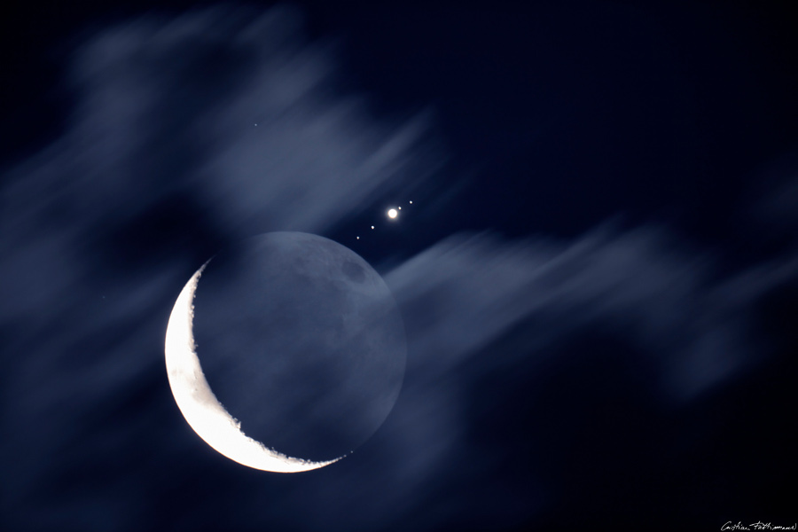 Hinter einem Schleier aus ziehenden Wolken leuchtet die Mondsichel, rechts davon ist der Jupiter mit seinen vier galileischen Monden.