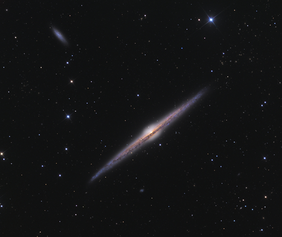 Eine spindelförmige Galaxie liegt diagonal mitten im Bild, umgeben von wenigen Sternen, die über den schwarzen Hintergrund versprenkelt sind. 
