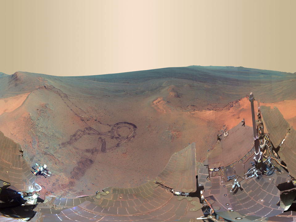 Das Panorama wurde waagrecht verkürzt. Links ist ein Kraterhang mit älteren Spuren des Rovers Opportunity. Unten ragen Solarpaneele ins Bild, die mit rötlichem Marsstaub bedeckt sind.