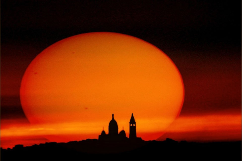 Am dunklen Himmel zeichnet sich die rot gefärbte Abendsonne bei Sonnenuntergang verzerrt ab. Als Silhouette steht unten die Basilica Sacré-Cœur de Montmartre in Paris.