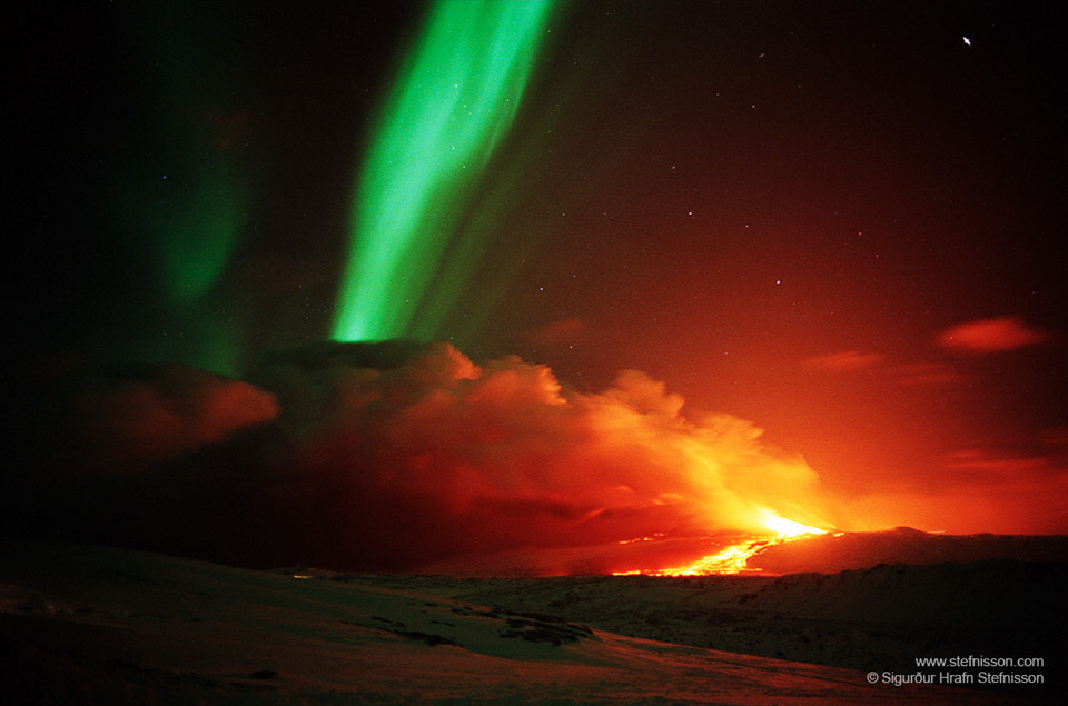 Am Himmel leuchtet ein grünes Polarlicht, es hat die Form eines schrägen Bandes. Rechts unten leuchtet glühende Lava im Dunkeln.