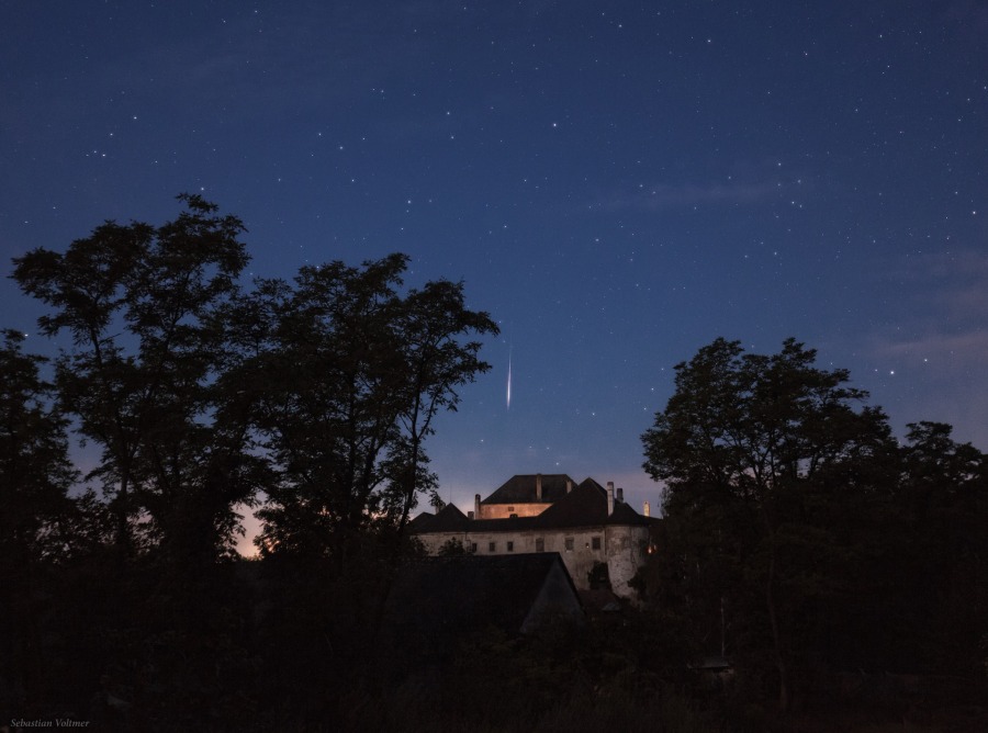 Über dem mittelalterlichen Schloss Albrechtsberg, das von den dunklen Silhouetten von Bäumen umgeben ist, blitzt am dunkelblauen Himmel ein Meteor der Perseïden auf und trifft scheinbar das Schloss.