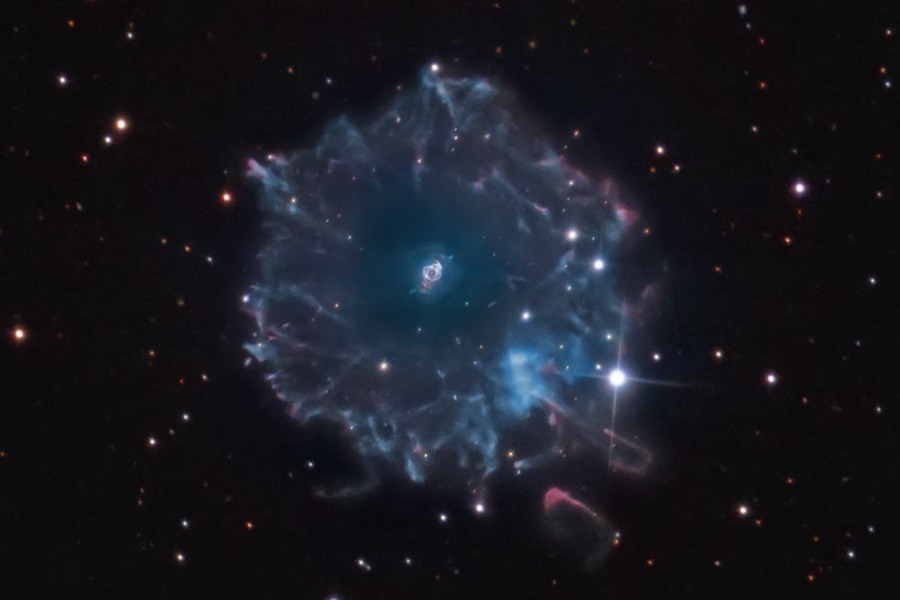 Der Katzenaugennebel NGC 6543 ist klein in der Mitte abgebildet. Außen herum verläuft ein riesiger, blasser Hof mit welligen Rändern.