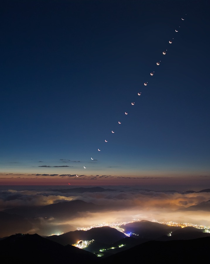 Über einer lichtverschmutzten Stadt, deren Helligkeit teilweise von Wolken abgemildert wird, steigt der Sichelmond auf und bedeckt dabei die Venus.