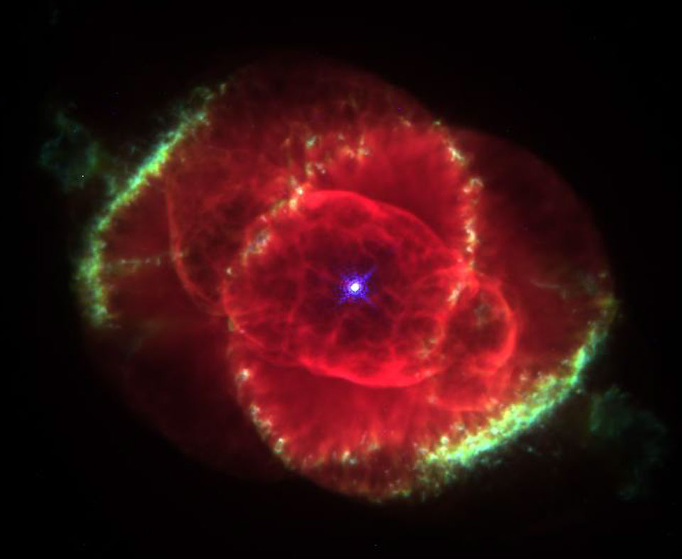 Das Hubble-Bild zeigt den Katzenaugennebel mit roten, komplexen Strukturen und zwei grünen Bögen am Rand. In der Mitte leuchtet ein heller Zentralstern.