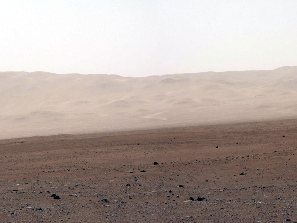 Das Bild zeigt die Aussicht des Marsrovers Curiosity. Hinter einer roten steinigen Marslandschaft ragt der Rand des Kraters Gale auf.