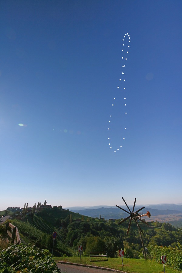 Über steirischen Weinbergen mit einem Klapotetz und einem Gebirge im Hintergrund wölbt sich am klaren blauen Himmel eine 8-förmige Schleife, die aus Einzelbildern der Sonne in leicht unregelmäßigen Abständen geformt wird.