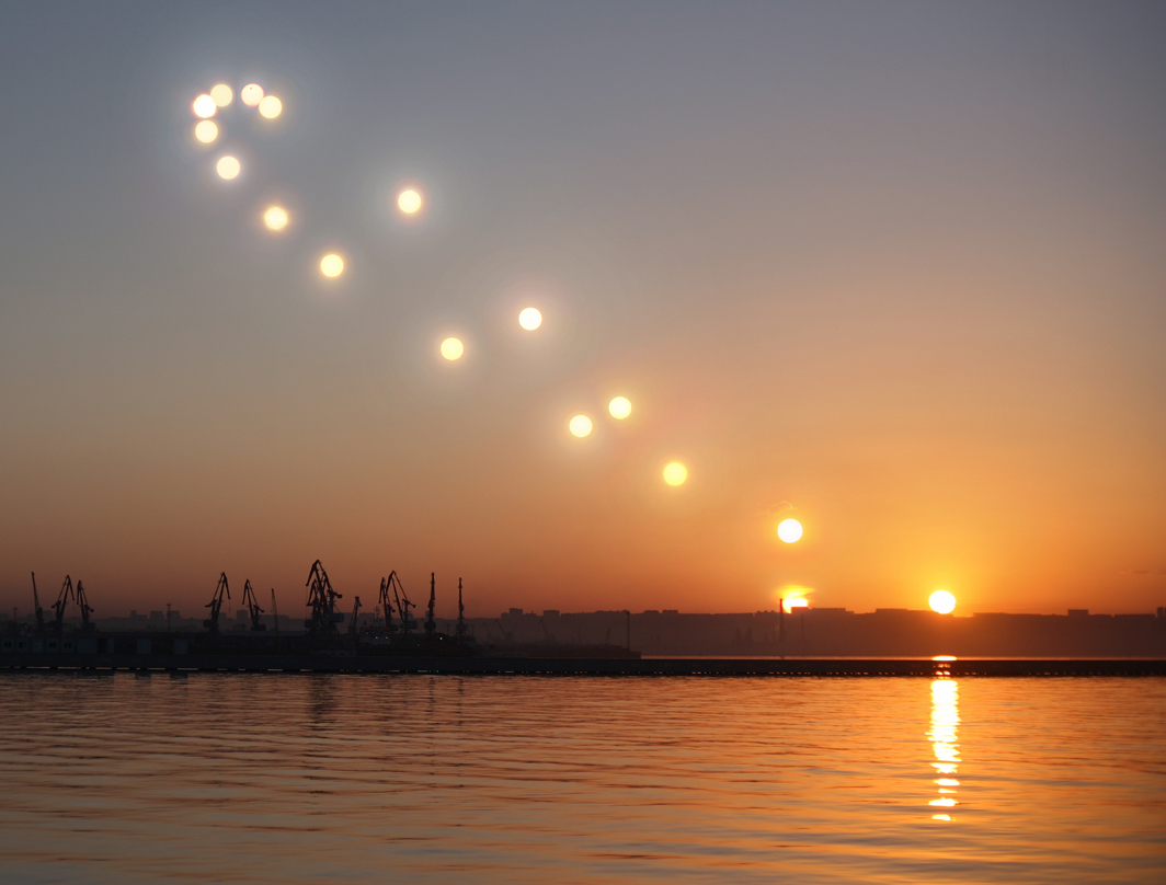 Über dem Kaspischen Meer verläuft im Osten der obere Teil eines Analemmas bei Sonnenaufgang. Auf einem Sonnenbild ganz oben ist ein kleiner Punkt. Es ist der Planet Venus während des Venustransits am 6. Juni.