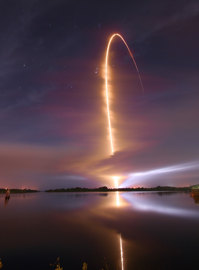 Hinter dem Turn Basin auf Cape Canaveral steigt eine bogenförmige Leuchtspur zum Himmel auf, sie spiegelt sich vorne im Wasser.