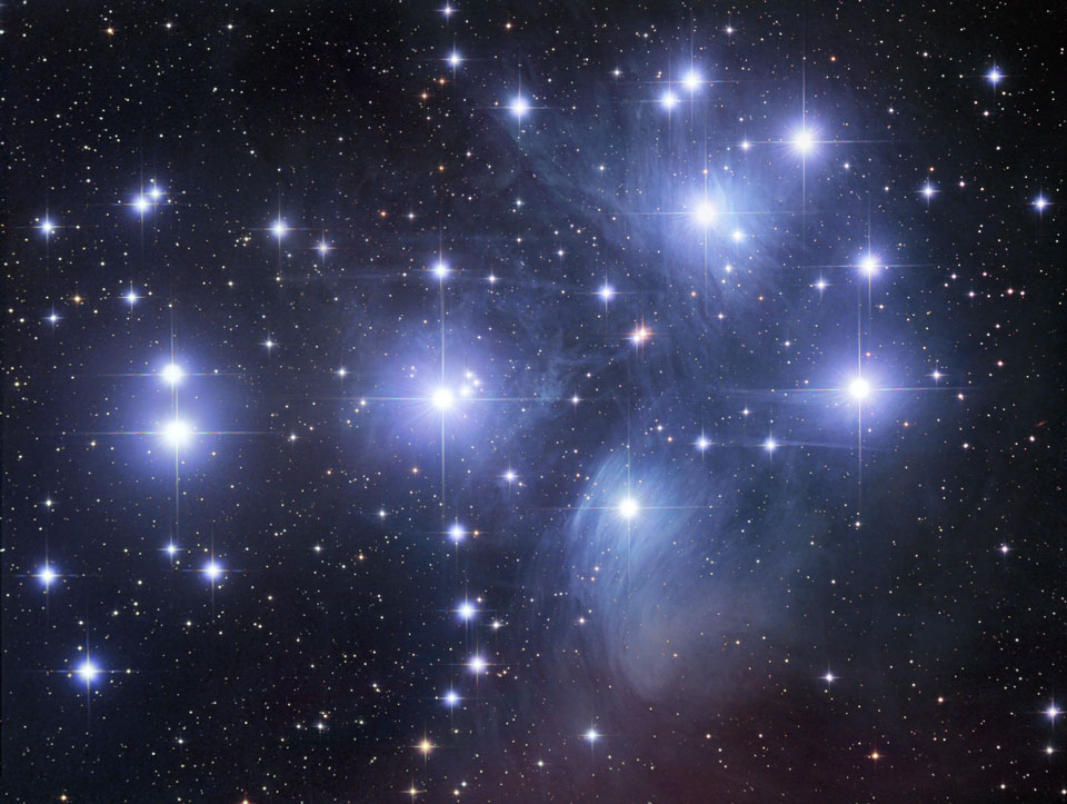 Die Sterne der Plejaden sind von blauen gefaserten Staubwolken umgeben. Die hellen Sterne sind von Lichtkreuzen umgeben, die durch den Fangspiegel im Teleskop entstehen.