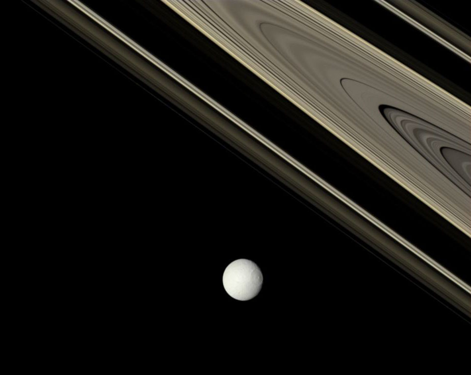 Rechts oben ragen die mächtigen Ringe Saturns markant ins Bild, darunter ist der grau-weiße Mond Tethys mit seinem riesigen Krater.