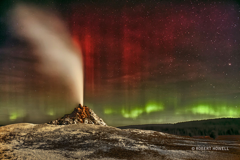 Aus einer Felsspalte strömt eine weiße Gasfahne. Dahinter schimmern grüne und rote Polarlichter, durch die Sterne zu sehen sind. Die Landschaft wird vom Mond beleuchtet.