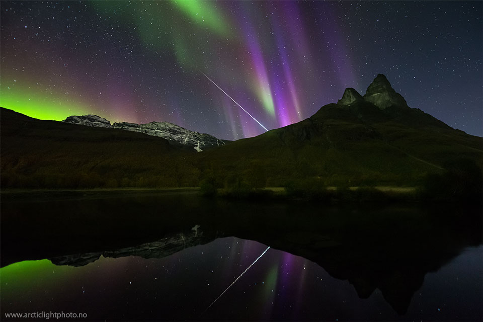 Hinter einem Berg, der sich vorne im Fluss Signalelva spiegelt, leuchten grüne und purpurfarbene Polarlichter, und eine Feuerkugel zischt schräg hinter den Berg.