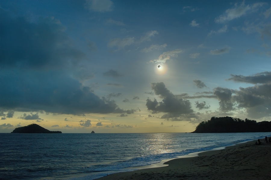 Am Himmel über dem Strand leuchtet die Korona, die eine totale Sonnenfinsternis mit Diamantring umgibt. Der Himmel ist noch von Wolkenfetzen überzogen.