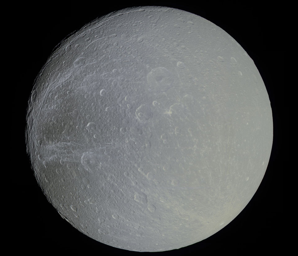 Der Saturnmond Dione wirkt eher grau als farbig. Seine Oberfläche wirkt sehr glatt mit flachen Kratern. Auf der Oberfläche sind auch Strahlen von Einschlägen zu sehen.