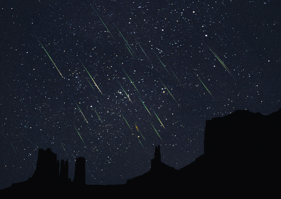 Hinter den Silhouetten der "Mittens", das sind Tafelberge im Monument Valley, strömen unzählige Meteore vor dem sternklaren Himmel zur Erde herab.
