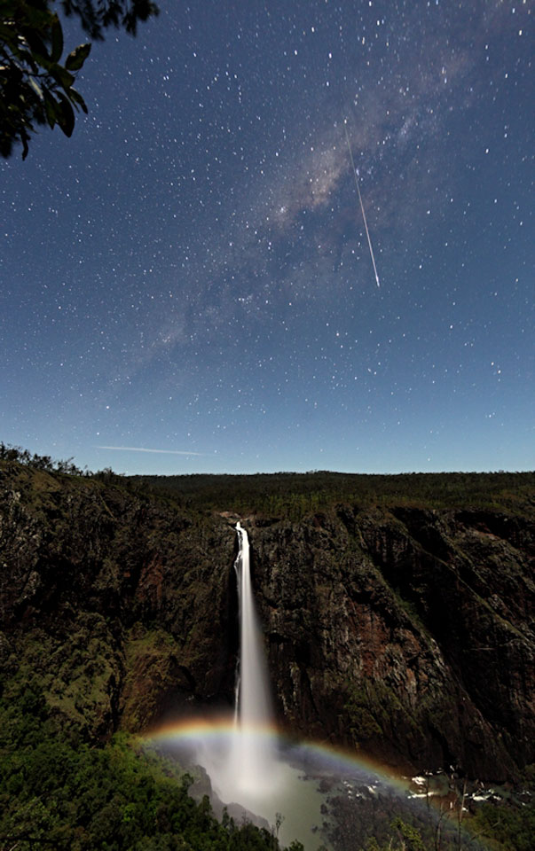 Von einer Klippe stürzt ein ruhiger Wasserfall in die Tiefe. Unten leuchtet ein bunter Mondregenbogen. Am Himmel blitzt ein Meteor vor dem Band der Milchstraße und dem sternklaren Himmel.
