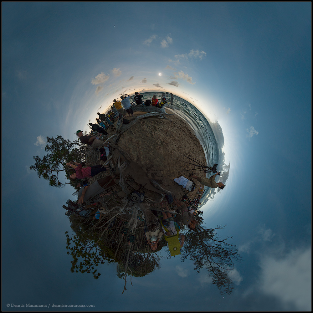Am blauen Himmel schwebt ein kleiner Planet mit einem Strand, über dem eine totale Sonnenfinsternis stattfindet. Das Bild entstand aus einem Panorama von Green Island an der Küste von Queensland.
