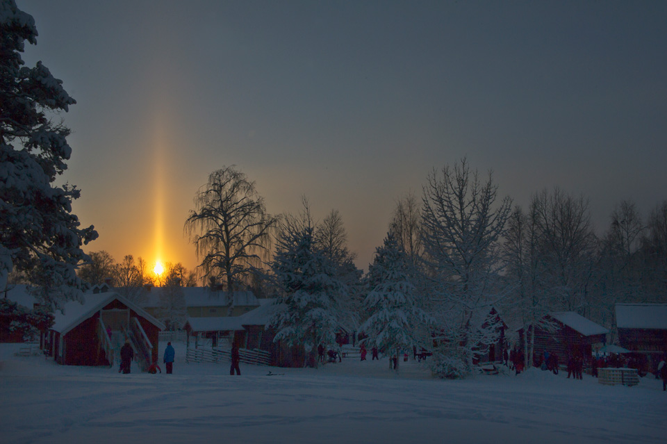 Hinter einer verschneiten Landschaft steigt eine helle Sonnensäule auf. Eiskristalle, die zu Boden flattern, erzeugen solche Lichtsäulen.