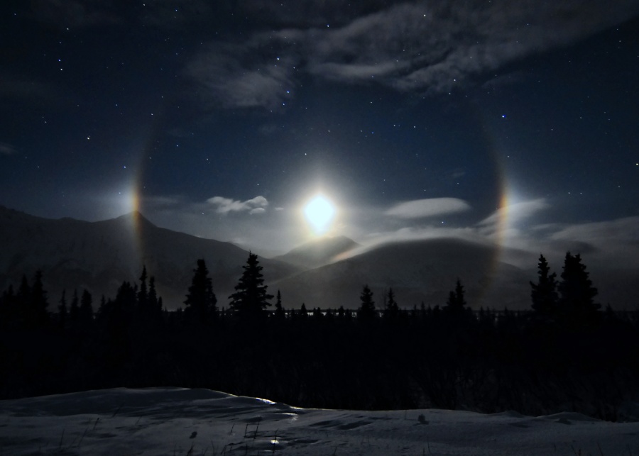 Der Mond leuchtet tief über einer gebirgigen Landschaft bei Lower Miller Creek in Alaska. Links und rechts neben dem Mond sind zwei Nebenmonde, sogenannte Paraselenae. Der Mond ist von einem blassen Halo umgeben.