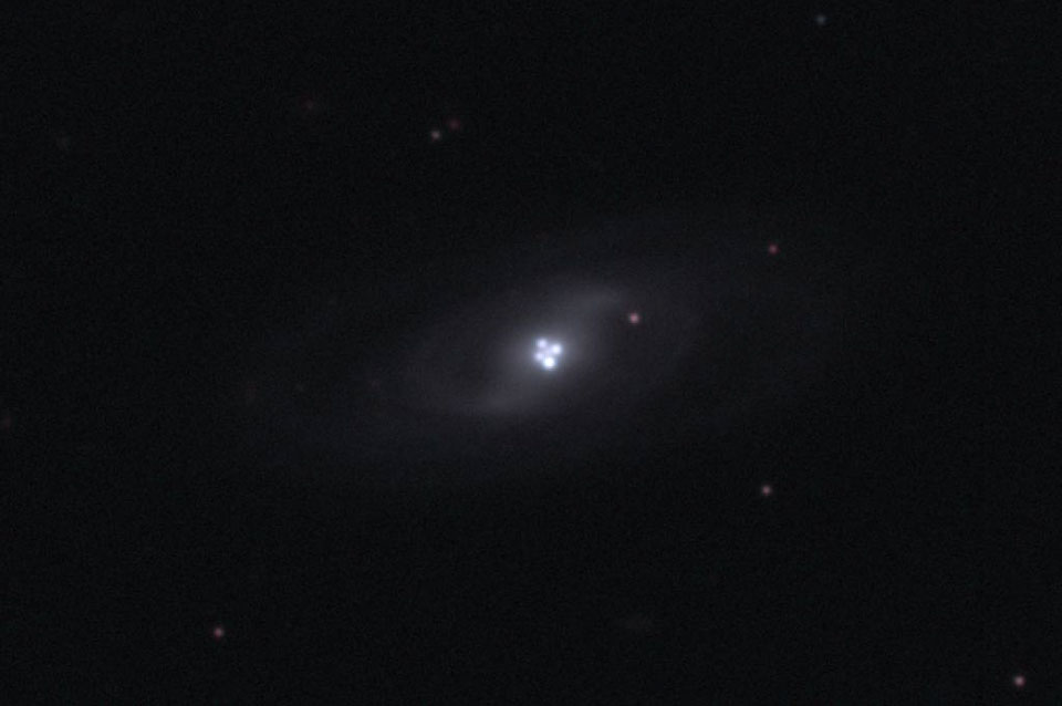 Mitten im Bild ist eine sehr blasse Galaxie mit Spiralarmen. In der Mitte leuchten sehr helle Flecken in Form eines Kleeblattes, sie bilden ein Einsteinkreuz und stammen von einem dahinter liegenden Quasar.