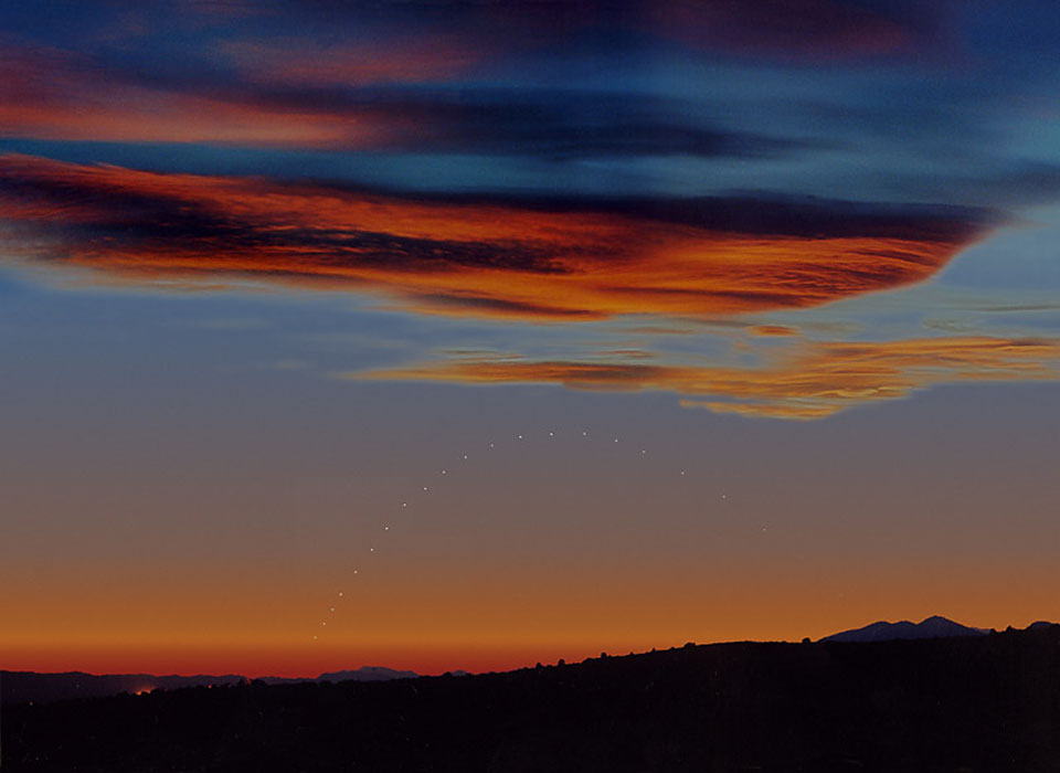 Über einem dunklen Horizont sind die Positionen des Planeten Merkur im März in der Abenddämmerung zu sehen. Wolken am klaren Himmel sind unten von Abendrot beleuchtet.