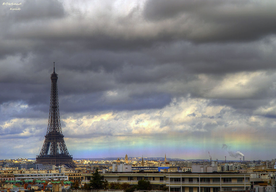Der Pariser Eiffelturm ragt über dem Horizont hoch. Darüber dräuen dunkle Wolken. Über dem Horizont verläuft waagrecht ein Horizontregenbogen.