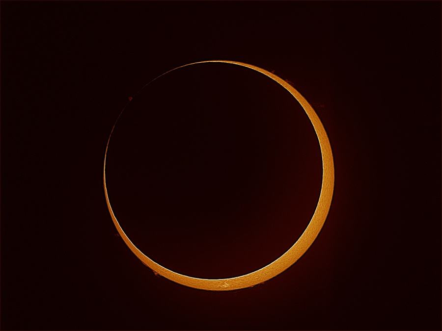 Am schwarzen Himmel prangt bildfüllend ein Feuerring. Die ringförmige Finsternis wurde mit H-alpha-Filter aufgenommen, daher ist die Struktur der Oberfläche erkennbar. Am unteren Rand ragt eine Protuberanz über den Sonnenrand.