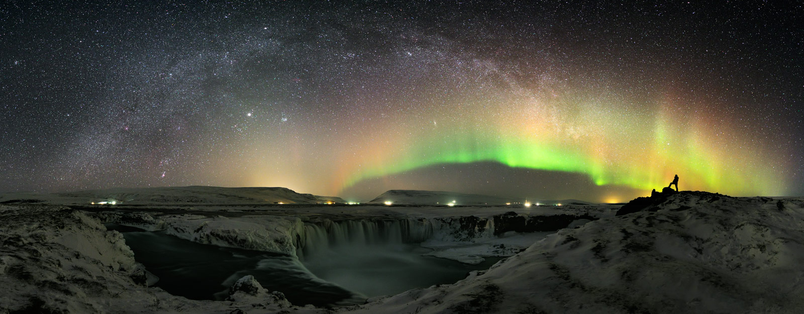 Über dem isländischen Wasserfall Goðafoss wölben sich ein grünes Polarlicht und die Milchstraße.Der Planet Jupiter und die Andromedagalaxi sind unter der Milchstraße zu erkennen.