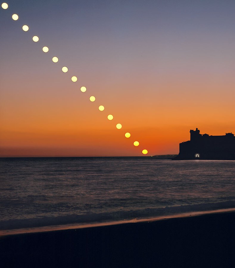 Über dem Tyrrhenischen Meer bei Santa Severa geht die Sonne unter. Rechts am Horizont steht die Silhouette einer Burg. Davor ist eine Figur erkennbar, die von einem Lichtschein umgeben ist. Es ist der Fotograf.