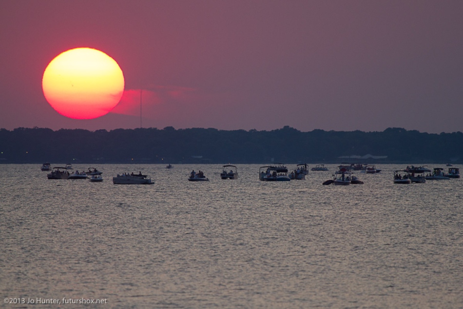 Auf dem Cedar Creek Lake südöstlich der texanischen Stadt Dallas schwimmen viele Schiffchen. Dahinter geht die Sonne unter. Sie ist in der Abenddämmerung gerötet und verzerrt. Mitten auf der Sonnenscheibe ist ein Sonnenfleck erkennbar.