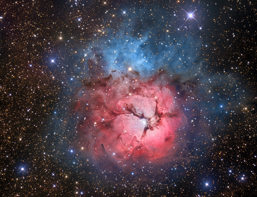 Der leuchtendrote Emissionsnebel in der Mitte ist von dunklen Staubranken dreigeteilt. Von oben her verläuft blauer Dunst eines Reflexionsnebels um die Emissionsregion. Das Bild zeigt den berühmten Trifidnebel im Sternbild Schütze.