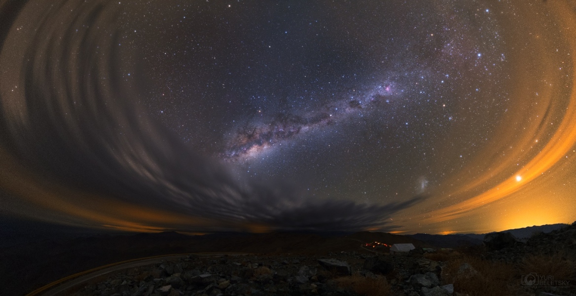 Rechts und links ziehen Sturmwolken schleifenförmige Gebilde am Himmel über der chilenischen Atacamawüste. Dazwischen steigt die Milchstraße schräg auf. Rechts unten leuchtet die Große Magellansche Wolke. Über orangefarbenen Stadtlichtern rechts unten strahlt der helle Stern Kanopus.