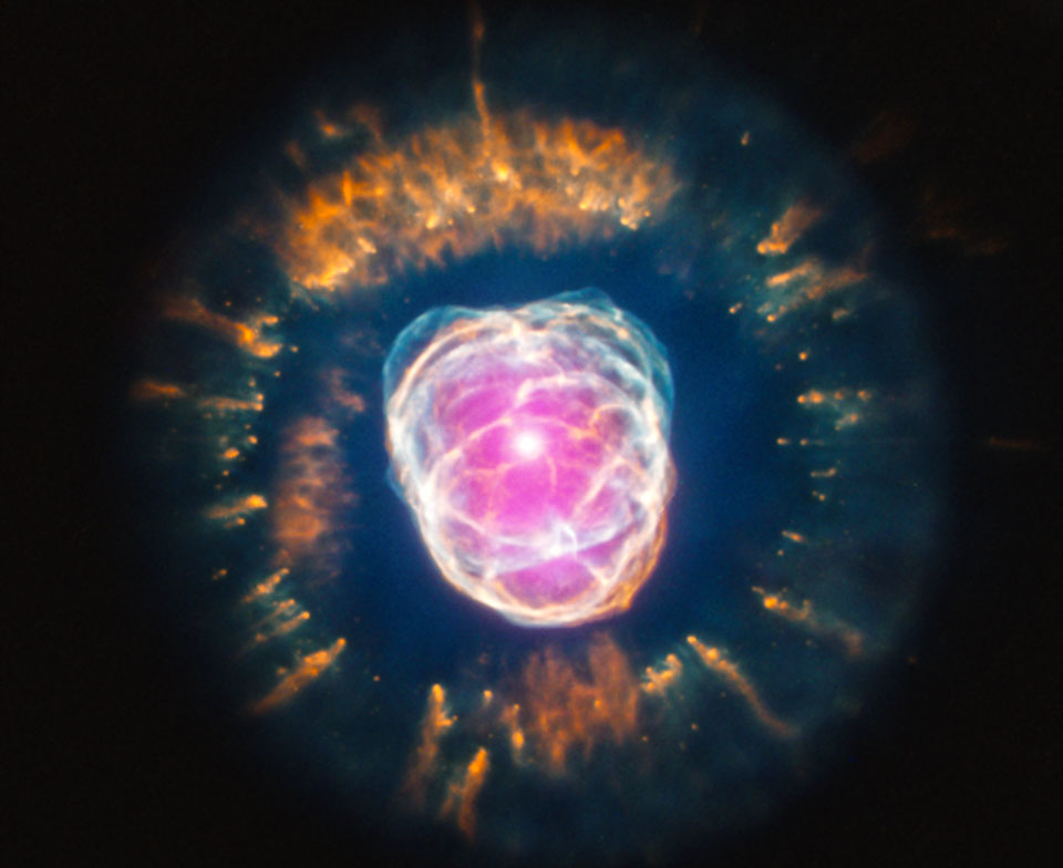 Das Innere dieses planetarischen Nebels leuchtet rosarot und ist von hellen Schlieren überzogen. Außen herum verläuft ein orangefarbener Strahlenkranz.