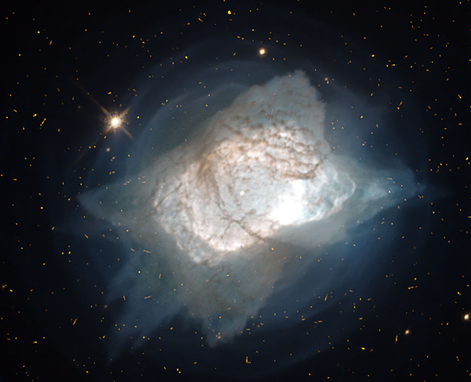 Der Nebel NGC 7027 erinnert ein wenig an ein Kissen oder an Popcorn. In der Mitte ist ein hell leuchtendes Oval, umgeben von einer Nebelschicht, außen herum ist ein schwach blaugrau leuchtender Hof.