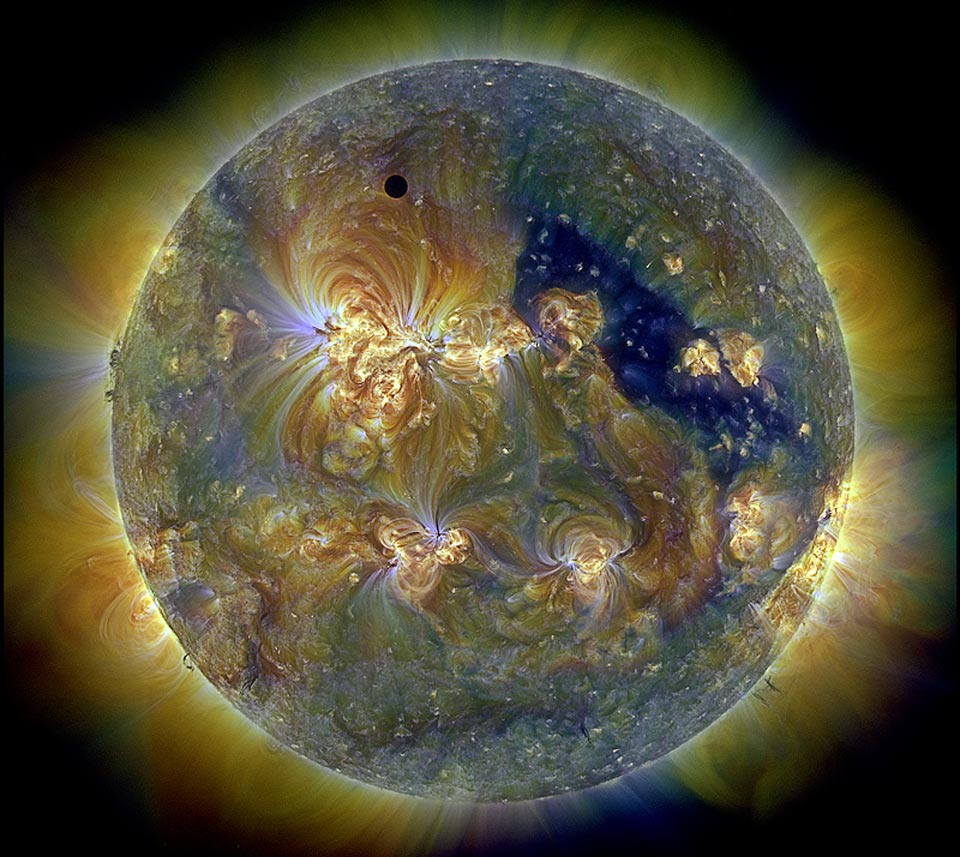 Das Bild zeigt die Sonne in drei Farben von Ultraviolettlicht. Links oben ist ein kreisrunder schwarzer Fleck, es ist der Planet Venus. Rechts ist ein dunkler Bereich, dort befindet sich ein koronales Loch.