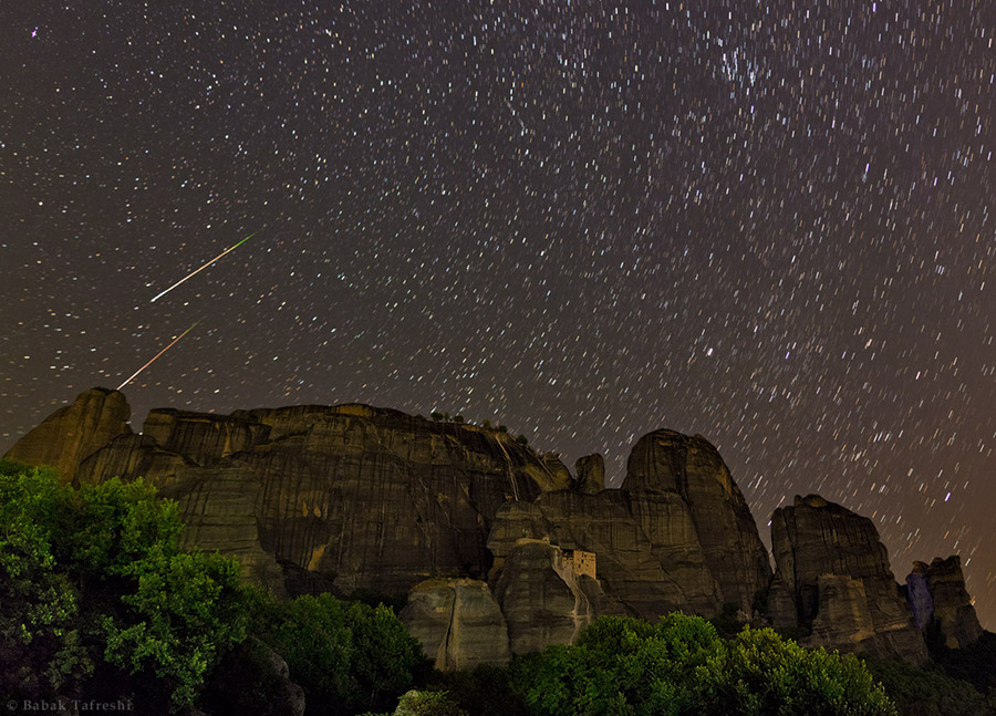 Auf den felsigen Klippen im Vordergrund steht das Kloster Meteora bei Kalabaka in Griechenland. Über den Himmel mit Strichspuren zischen zwei Meteore, rechts ist das Sternbild Perseus, in dem der Radiant der Perseïden liegt.
