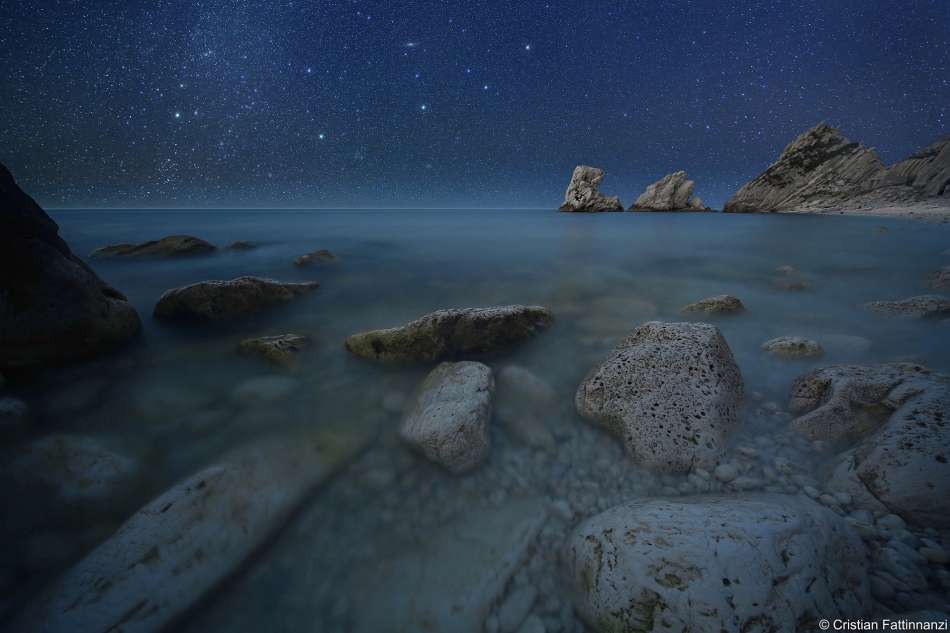 Hinter dem felsigen Ufer am Meer ist über dem Horizont der Sternenhimmel mit der Andromedagalaxie zu sehen.