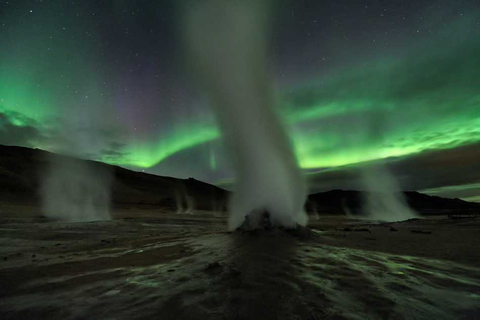 Aus einer nächtlichen Landschaft, die von grünen Polarlichtern beleuchtet wird, steigen Dampfsäulen auf.
