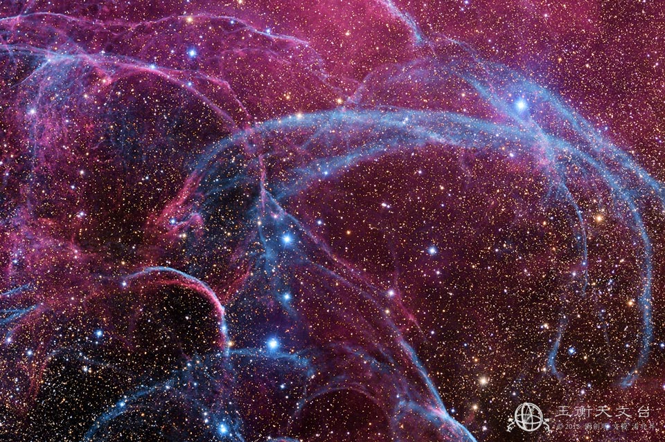 Das Bild ist dicht mit Sternen gesprenkelt. Dazwischen verlaufen Bögen, die blau und magentafarben leuchten. Einige Sterne sind heller und heben sich vom Sternenteppich im Hintergrund ab.