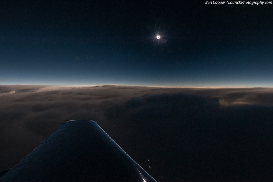 Ein Flugzeugflügel schwebt über den Wolken. Oben strahlt die Sonnenkorona um den Mond herum am dunkelblauen, fast schwarzen Himmel.