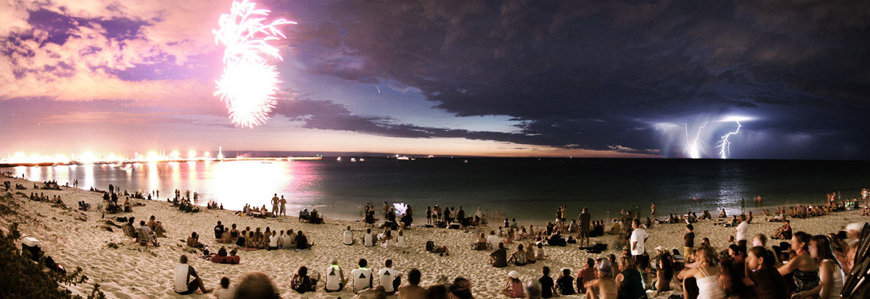 Am Strand sitzen viele Einwohner der australischen Stadt Perth und bewundern das Feuerwerk zum Australia Day. Rechts leuchten Blitze eines fernen Gewitters. In der Mitte steht in einer Wolkenlücke der Komet McNaught mit seinen prachtvollen Schweifen.